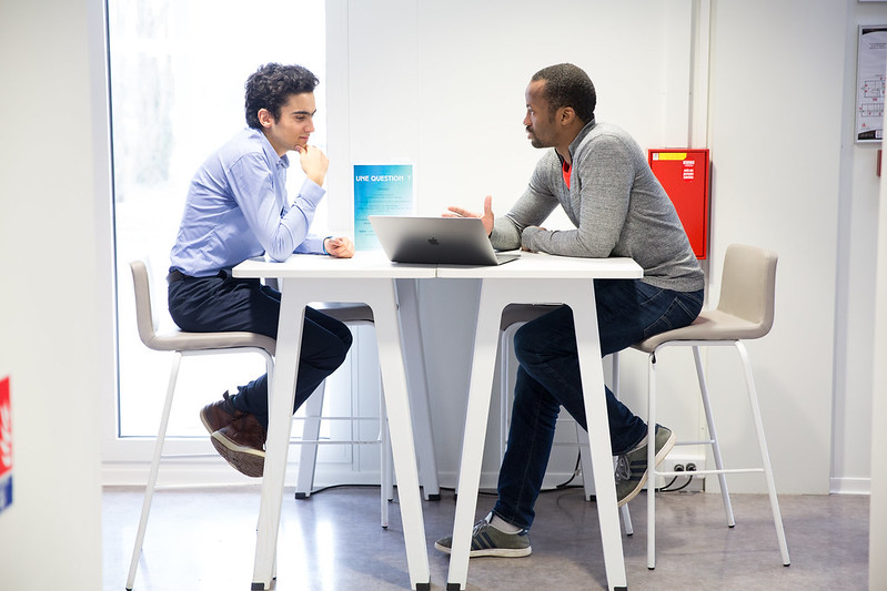 Deux hommes s'assoient à une table avec un laptop entre eux. Ils parlent.