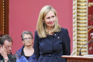 Monique Leroux, présidente du conseil d'Investissement Québec, se tient débout à gauche d'un podium. Il y a deux autres gens derrière d'elle. Elle a un petit sourit et écoute le speaker au podium.