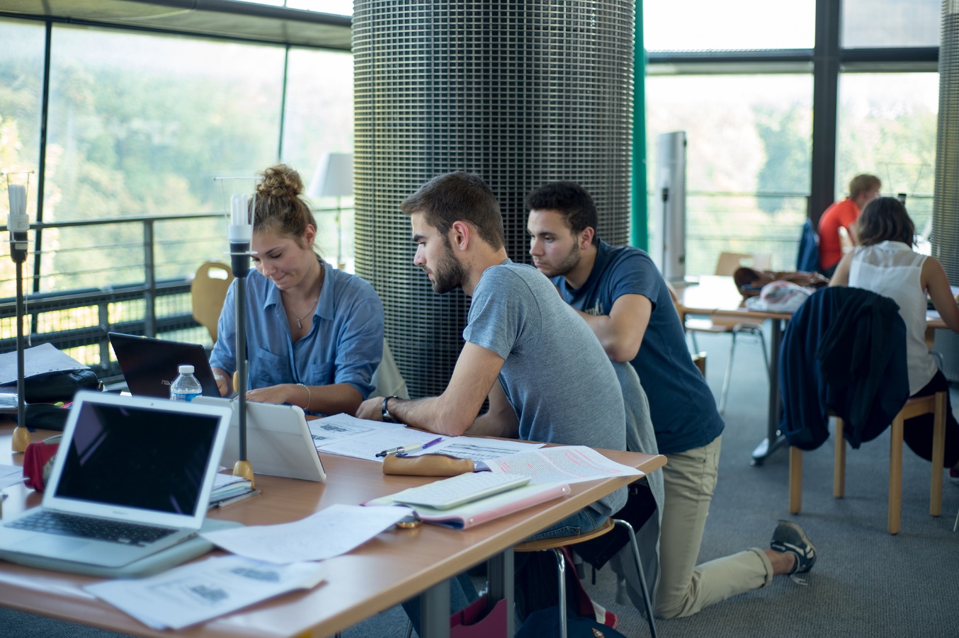 Trois étudiants travaille ensemble dans une salle bien ensoleillée. Il y a une jeune femme et un jeune homme s’assoient à une table couverte avec des papiers et des laptops, et il y a un autre homme à genoux à coté de la table.